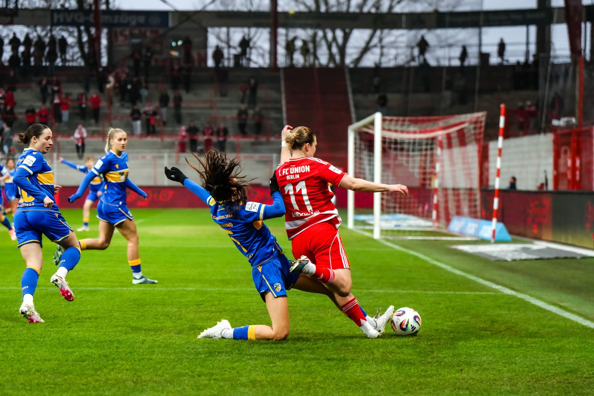 Spielszene im Fußball: Eine Spielerin dribbelt, während eine gegnerische Spielerin sie im Tackle zu Fall bringt. Zuschauer im Hintergrund.