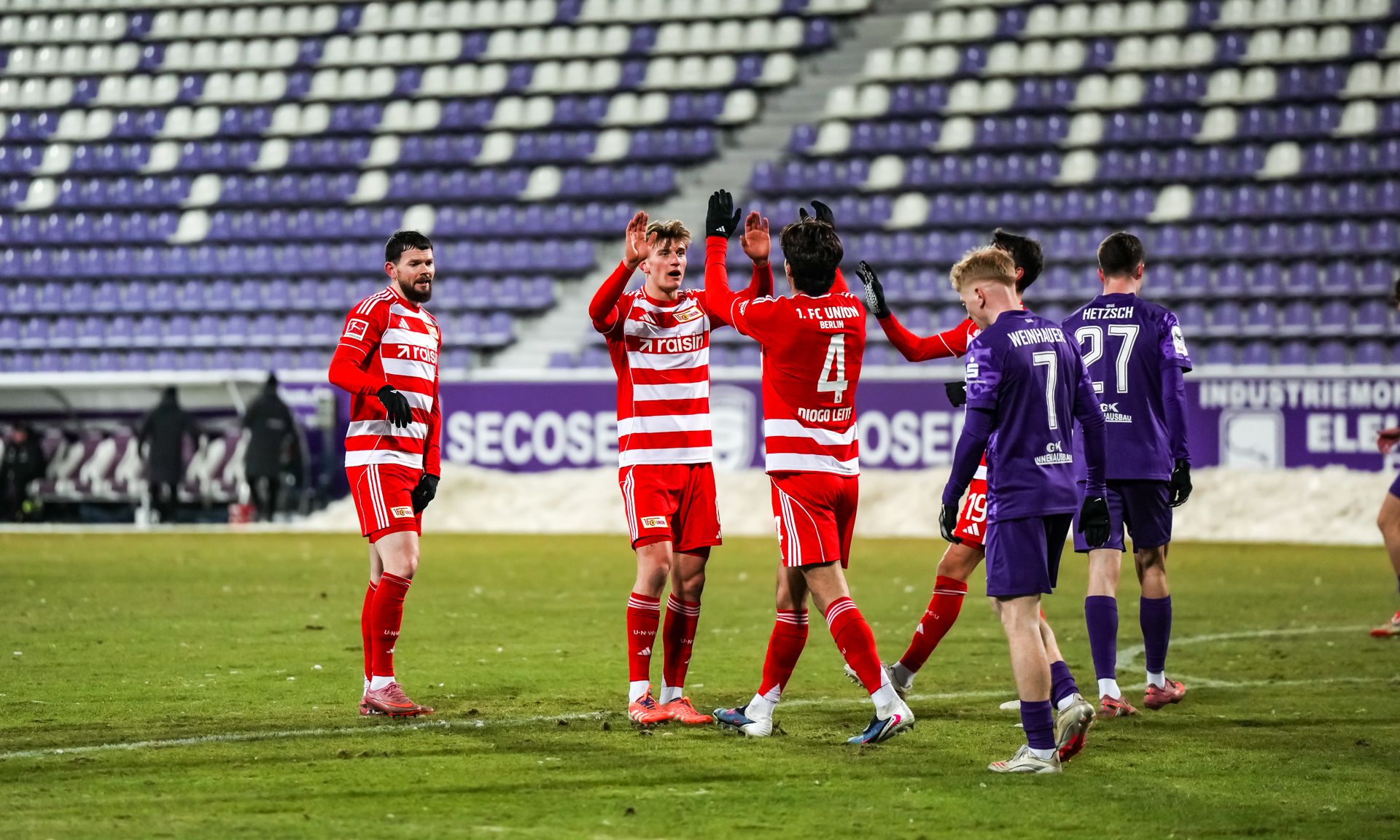 Spieler in roten Trikots feiern zusammen auf dem Spielfeld, während Gegner in lila Trikots zuschauen. Stadion mit leeren Rängen im Hintergrund.