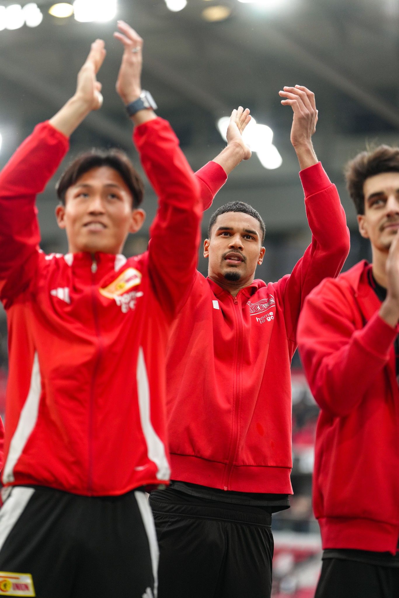 Drei Fußballspieler in roten Trikots applaudieren in einem Stadion.