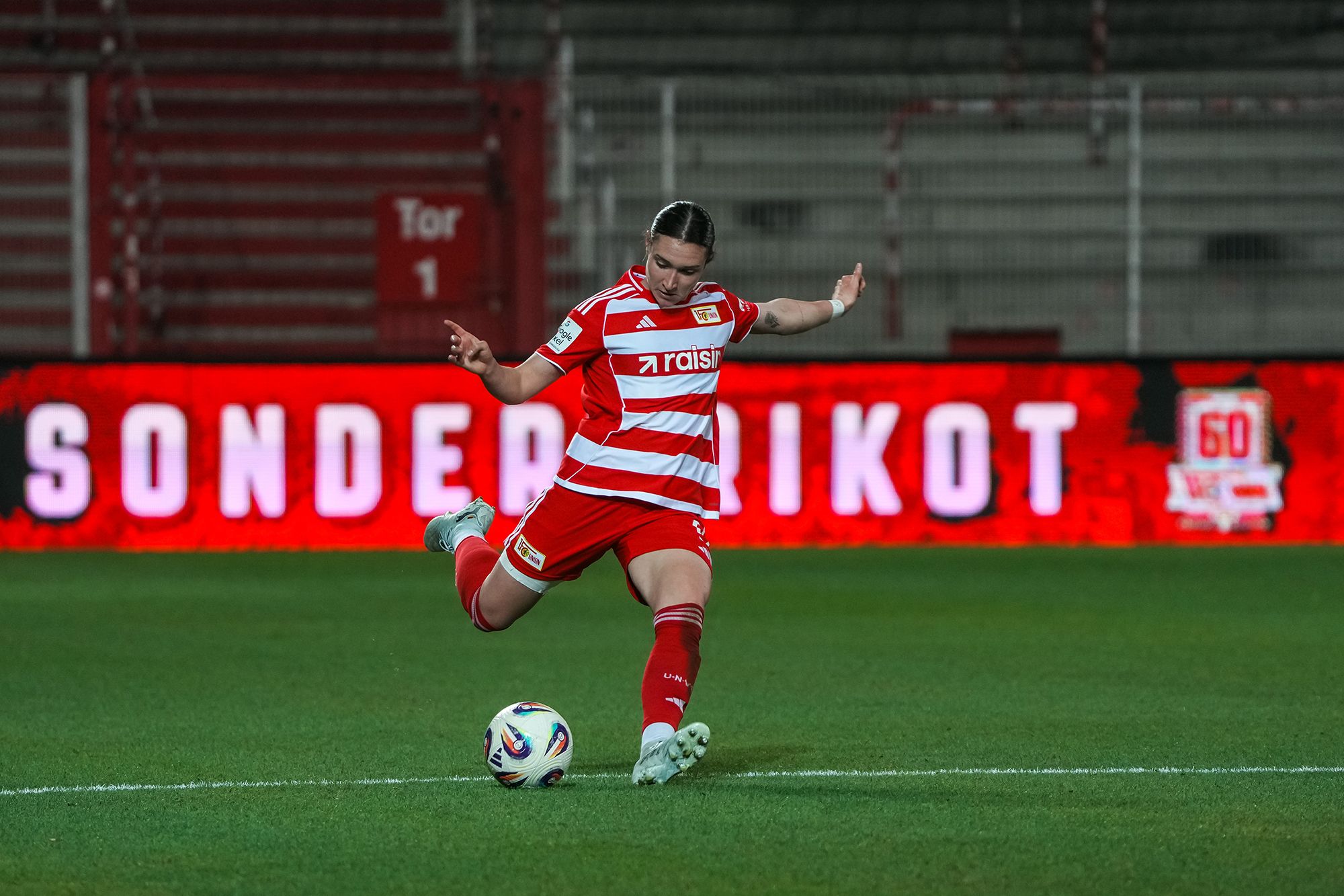 Eine Fußballspielerin in rot-gestreiftem Trikot führt einen Schuss aus, im Hintergrund leuchtet eine Werbebande.