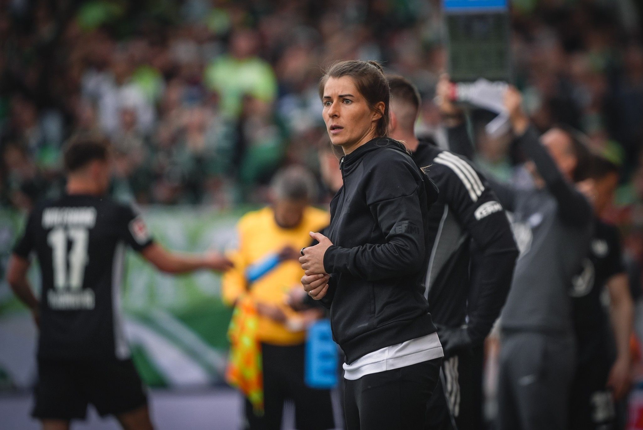 Eine Trainerin steht am Spielfeldrand und beobachtet das Spiel, während andere Teammitglieder im Hintergrund stehen.