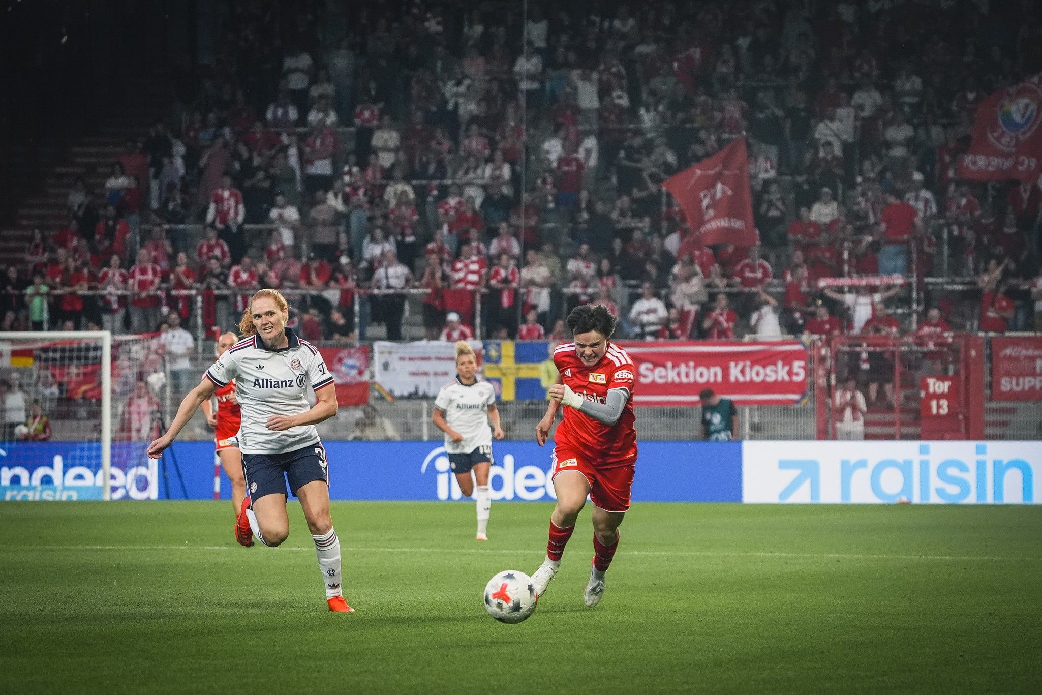 Ein Fußballspiel mit Spielern in roten und blauen Trikots, umgeben von jubelnden Fans im Stadion.