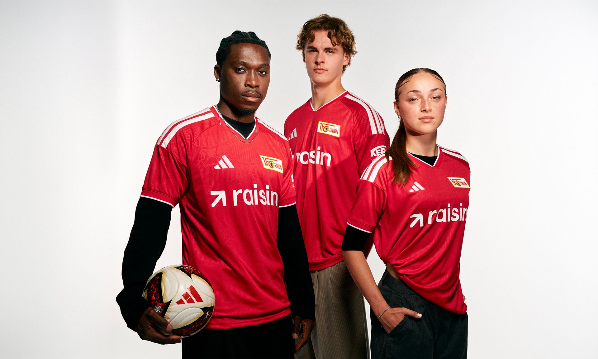 Drei Sportler in roten Fußballtrikots mit Sponsorenlogo, einer mit Ball, vor neutralem Hintergrund.