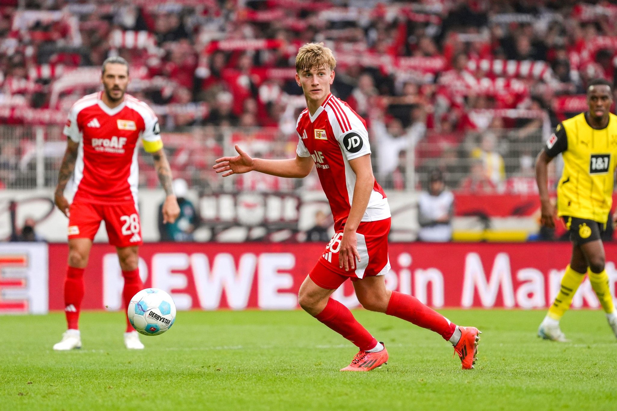 Ein Fußballspieler in rotem Trikot läuft mit dem Ball auf dem Spielfeld, unterstützt von jubelnden Fans im Hintergrund.