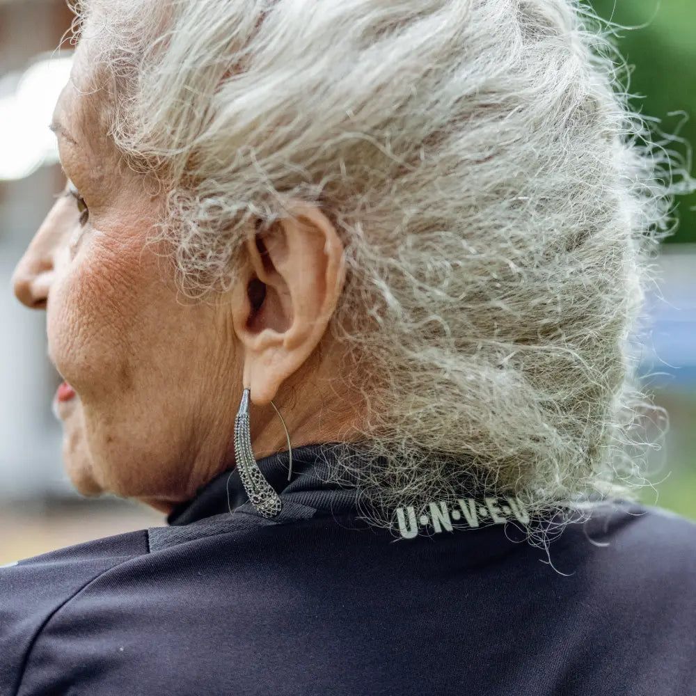 Eine ältere Frau hat graue, lockige Haare. Sie trägt ein schwarzes Oberteil und einen großen Ohrring.