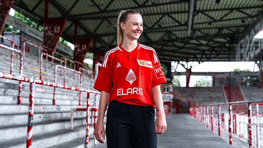 A smiling woman in a red soccer jersey stands in an empty stadium with metal railings.