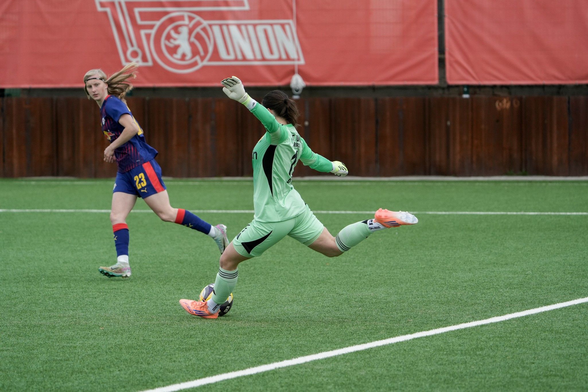 Eine Torhüterin in grünem Trikot sprintet nach dem Ball, während eine Spielerin in blau und rot heranläuft.