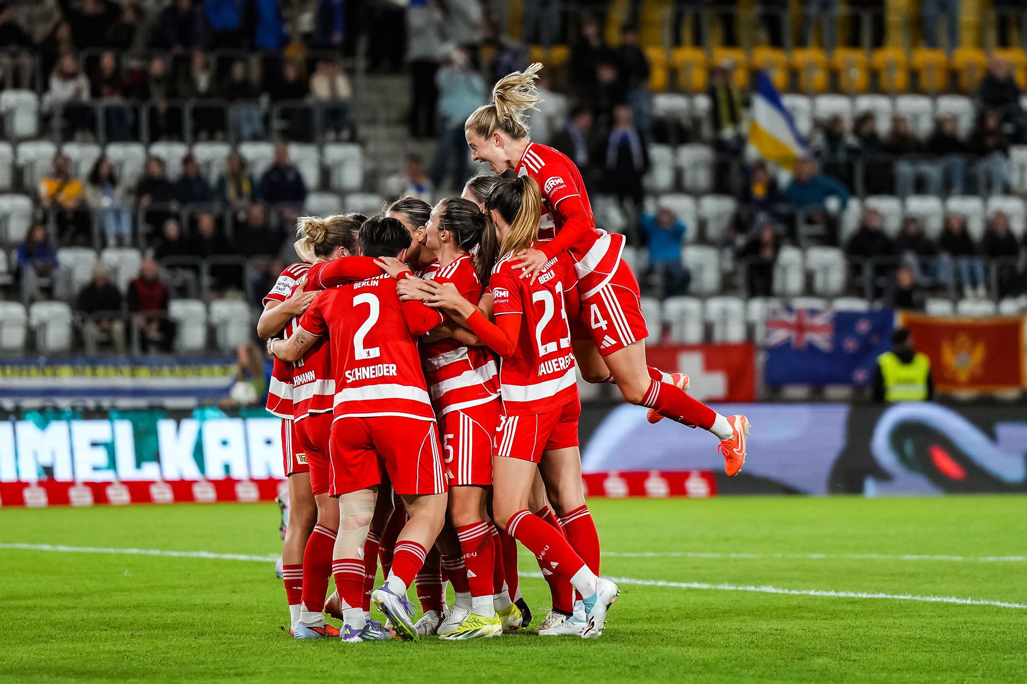 Ein Team von Frauenfußballspielerinnen in roten Trikots feiert einen Torerfolg auf dem Spielfeld.