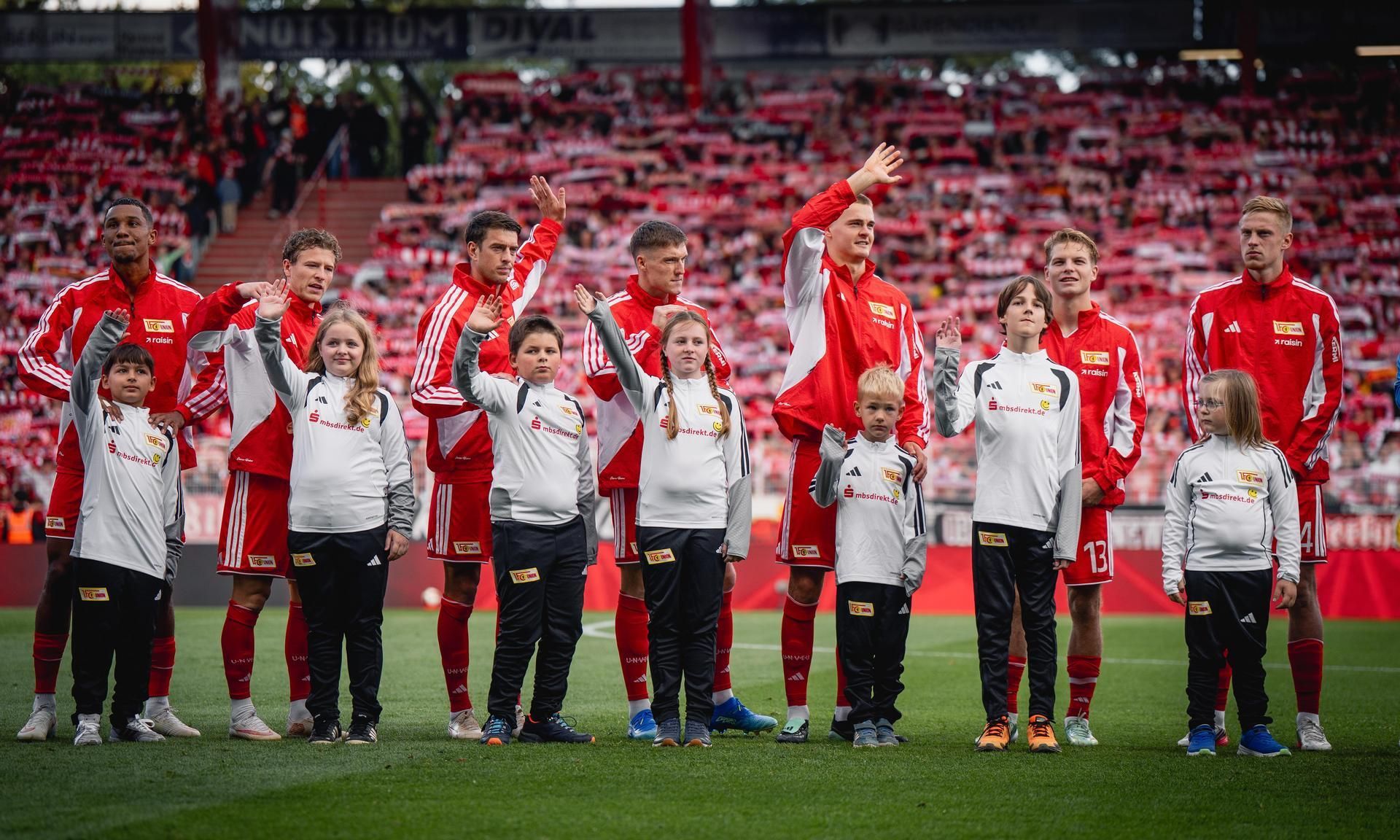 Spieler von Union Berlin stehen mit Kindern auf dem Spielfeld und winken in die Zuschauermenge.