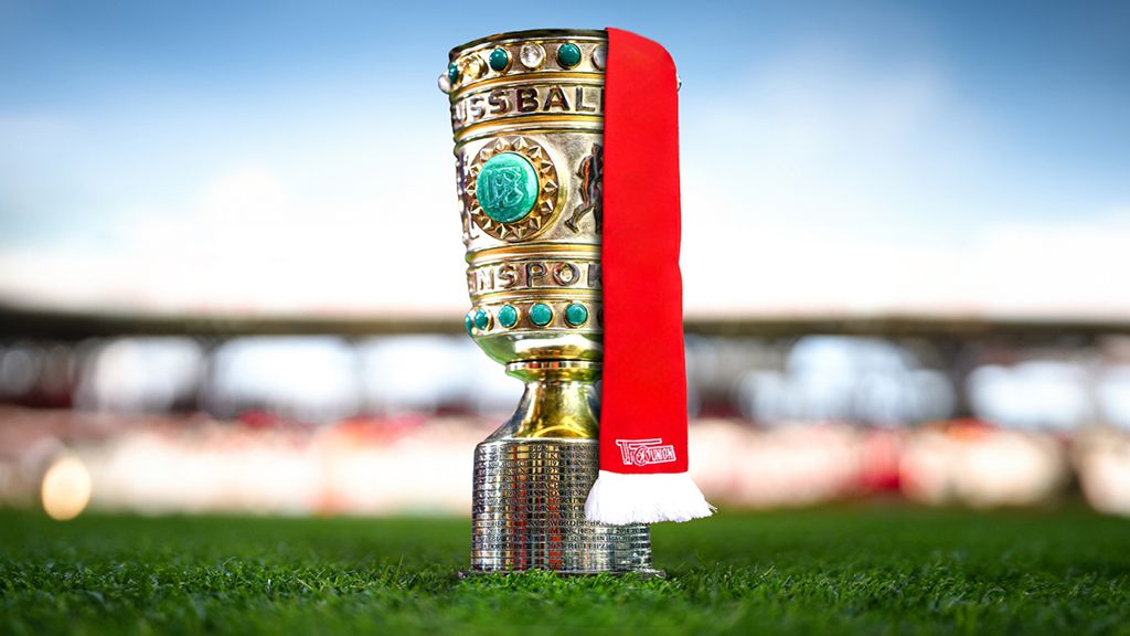 Ein goldener Fußballpokal steht auf einem grünen Rasenfeld, mit einer roten Schleife am Griff. Hintergrund ist unscharf.