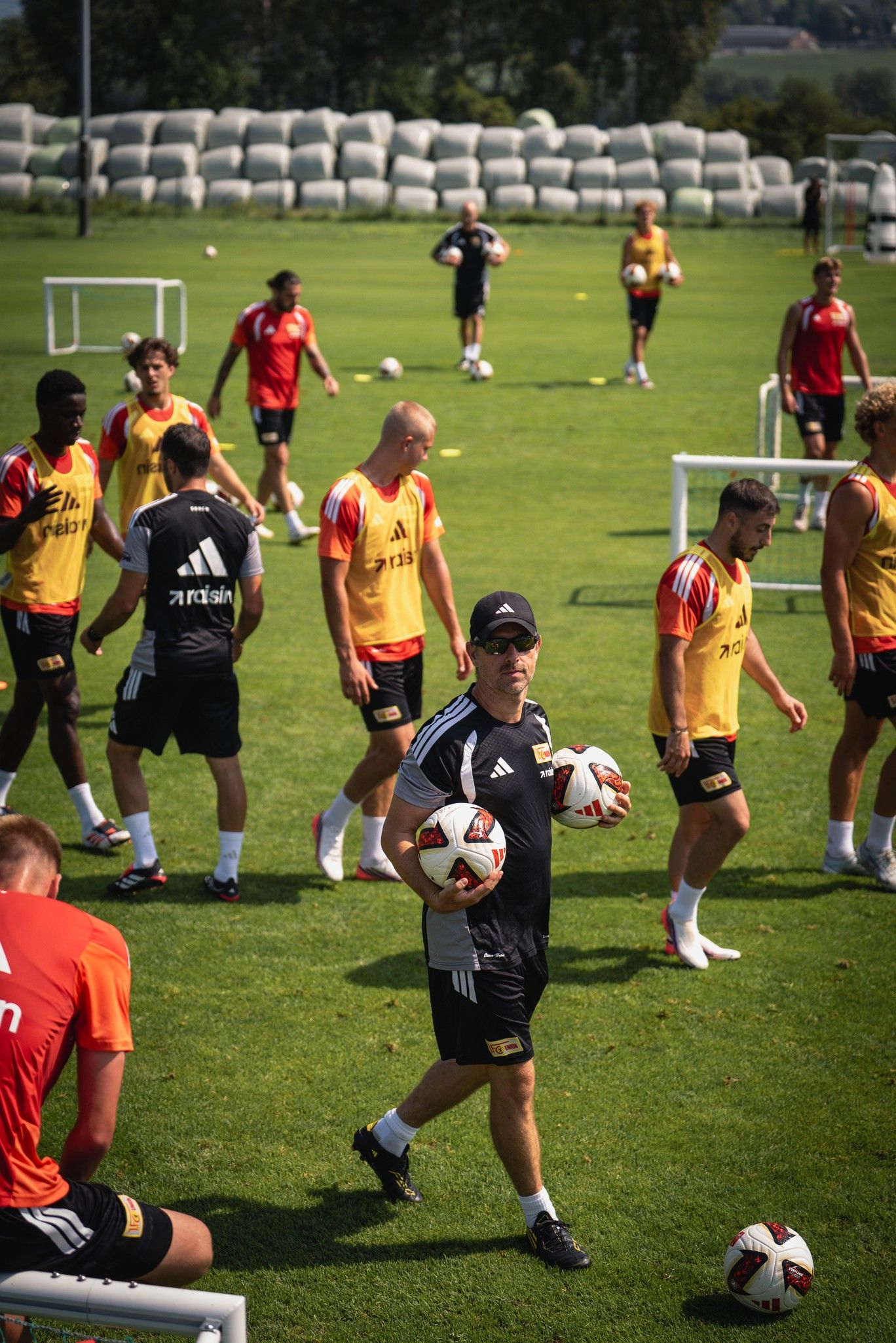 Trainer mit Bällen in der Hand, während im Hintergrund mehrere Spieler auf einem Sportplatz trainieren.