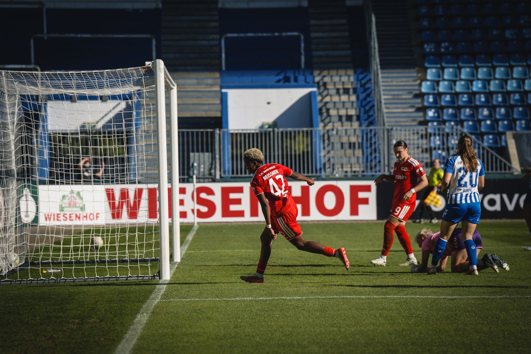 Spieler in roter Uniform jubelt nach einem Tor, während ein Torwart und ein weiterer Spieler in blauer Uniform am Boden liegen.
