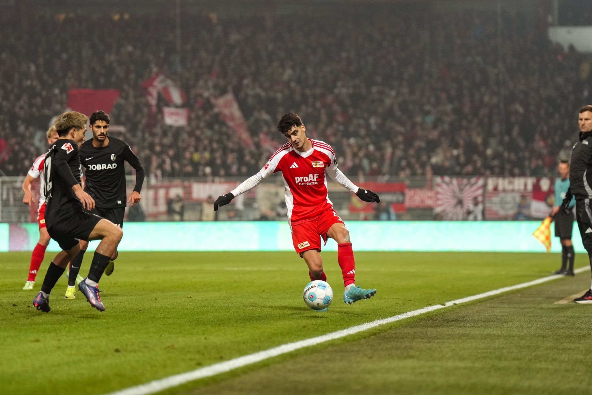 Game scene from a soccer match: A player in red uniform with the ball, surrounded by opponents and a cheering crowd in the background.