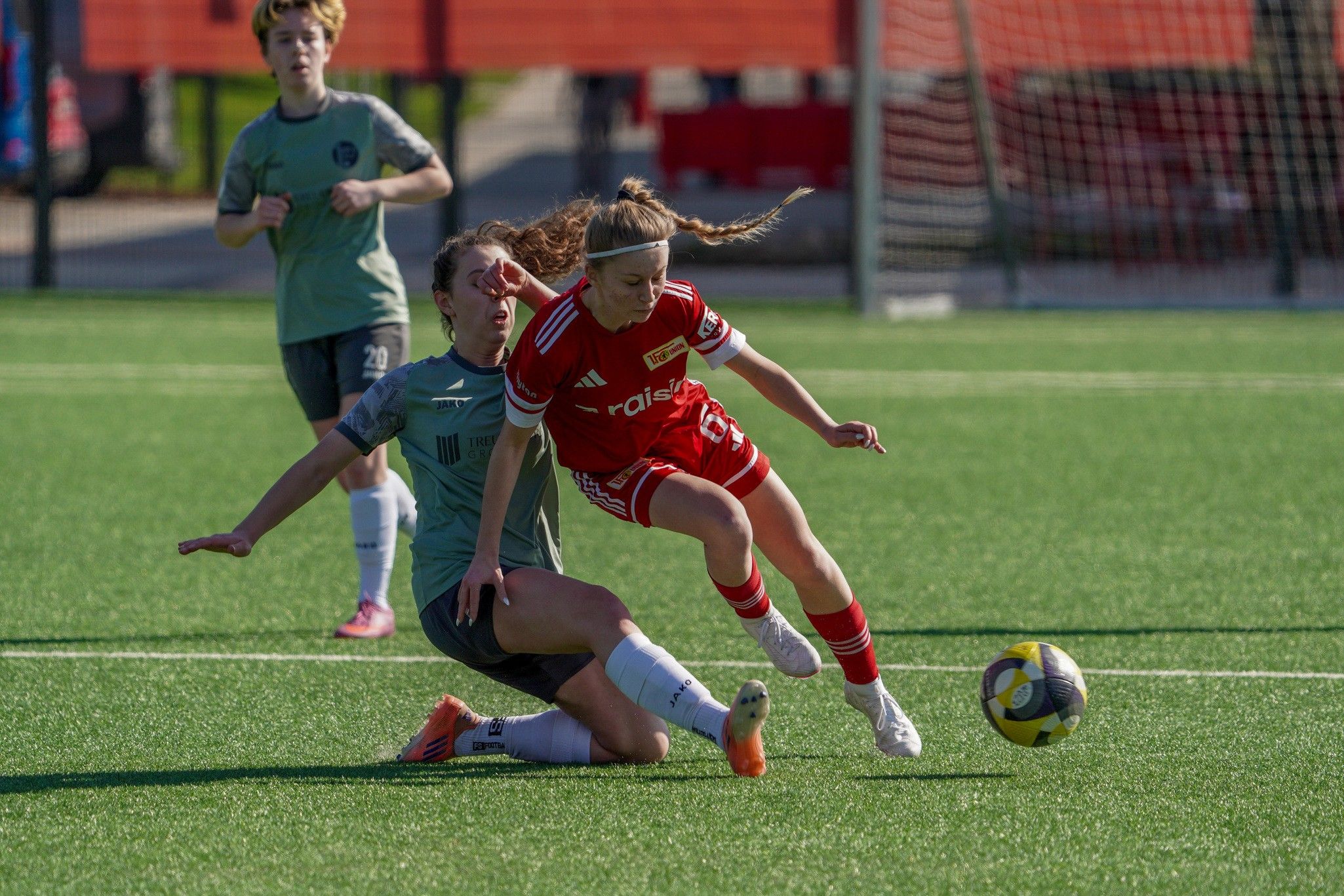 Eine Fußballspielerin in rot fällt während eines Spiels auf dem Rasen, während eine Gegenspielerin in Grau sie tackelt.