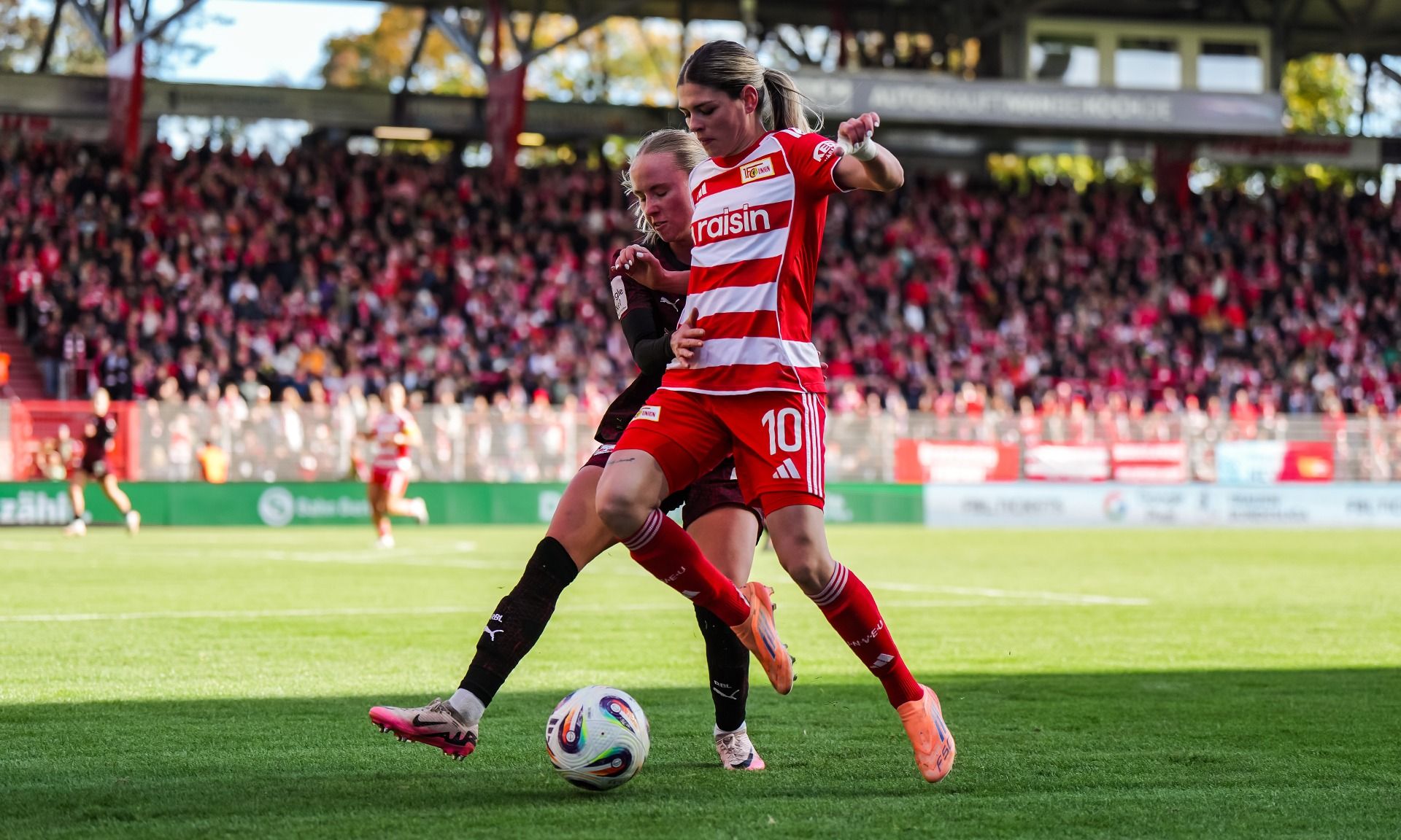 Zwei Spielerinnen im Fußballspiel kämpfen um den Ball auf einem Sportplatz vor einer jubelnden Menge.