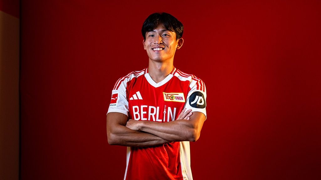 Lächelnder junger Fußballspieler in einem rot-weißen Trikot mit "Berlin"-Aufdruck auf rotem Hintergrund.