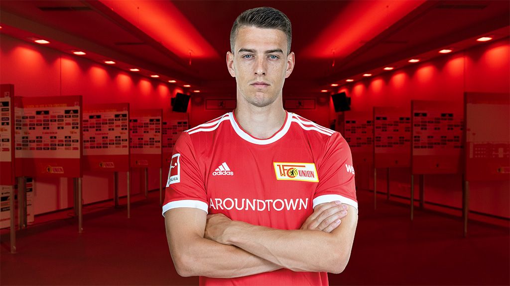 Ein junger Fußballspieler in einem roten Vereins-Trikot steht mit verschränkten Armen vor einer beleuchteten, roten Hintergrundkulisse.