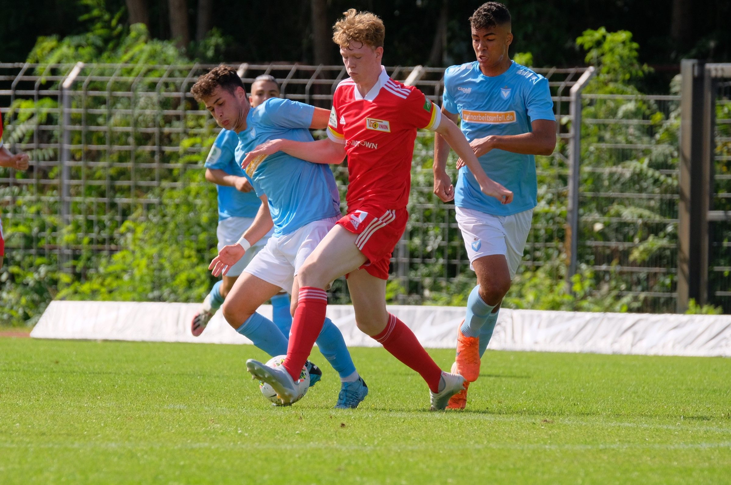 Junge Fußballspieler in rot und blau kämpfen um den Ball auf einem grünen Rasenplatz.