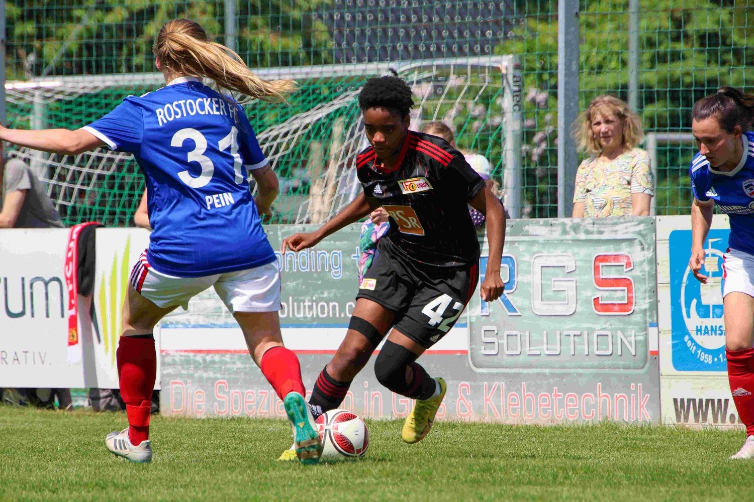 Zwei Fußballspielerinnen im Wettkampf auf einem Sportfeld, eine Spielerin trägt ein schwarzes Trikot, die andere ein blaues.
