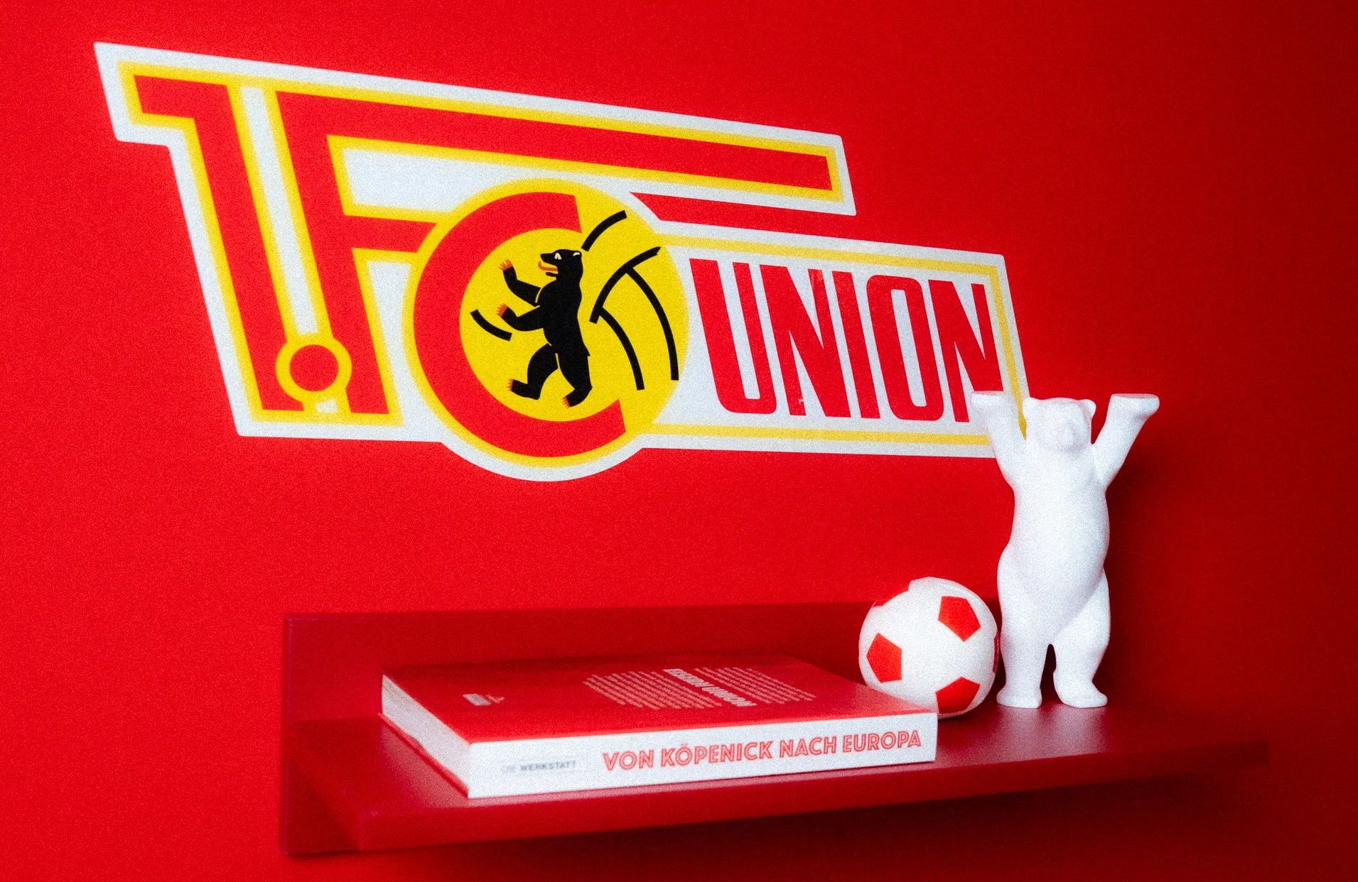 Wand mit dem Logo von 1. FC Union Berlin, einer weißen Bärenfigur und einem roten Fußball auf einem Regal.