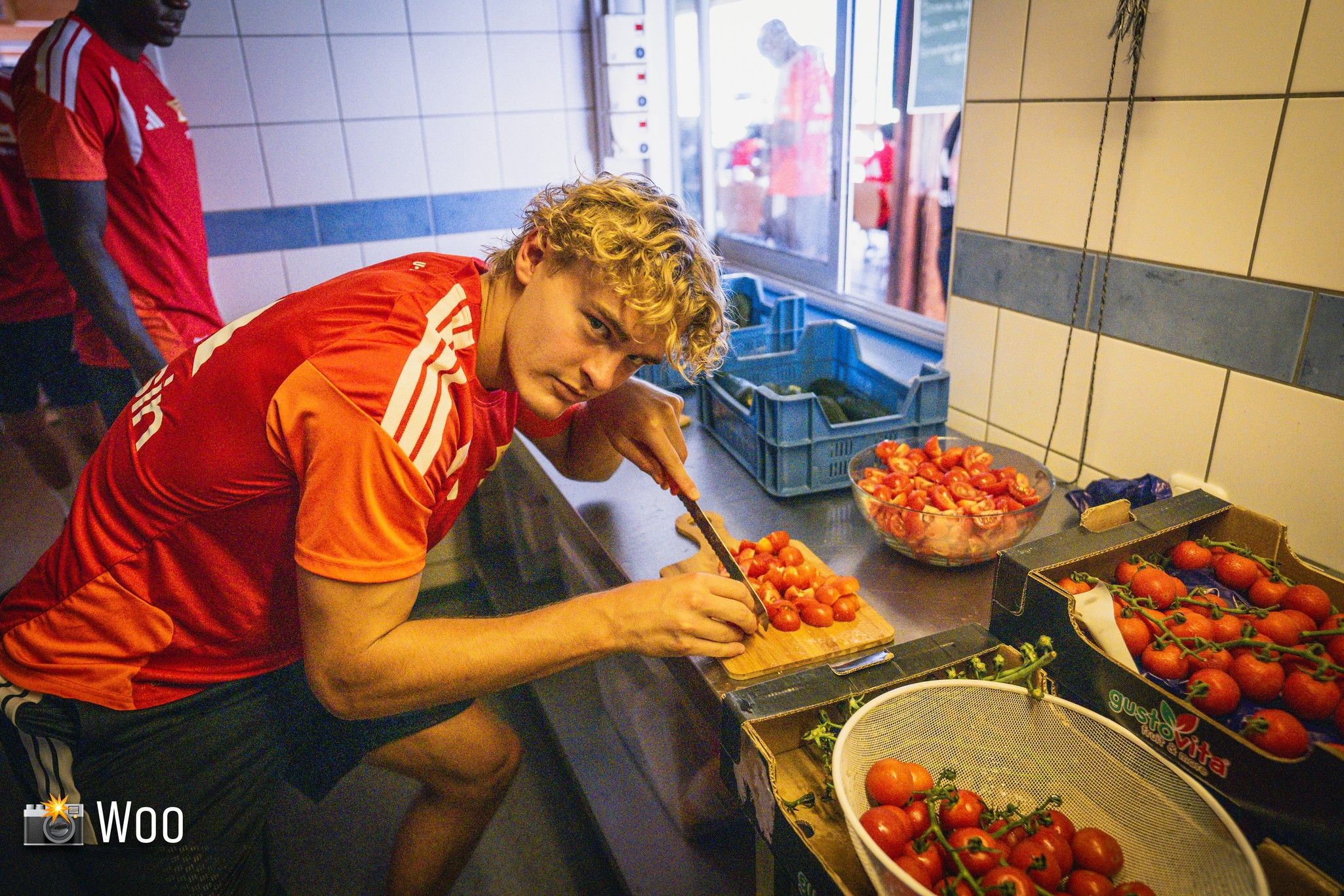 Ein junger Mann mit lockigem Haar schneidet Tomaten in einer Küche, während andere Personen im Hintergrund beschäftigt sind.
