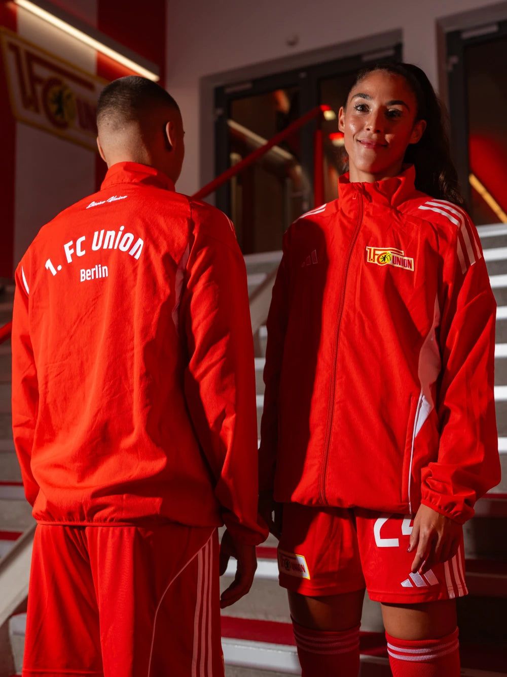 Zwei Athleten in roten Trainingsanzügen des 1. FC Union Berlin stehen in einem sportlichen Umfeld.