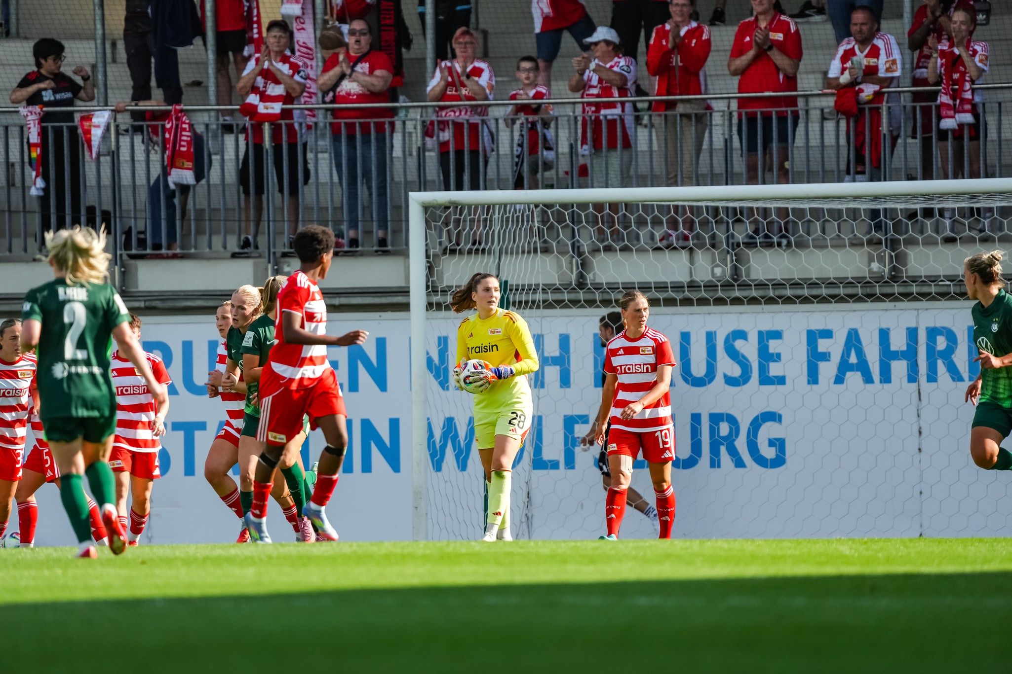 Fußballspiel mit Spielerinnen in rot-weiß gestreiften Trikots und einem Torwart in gelber Kleidung, umgeben von jubelnden Fans.