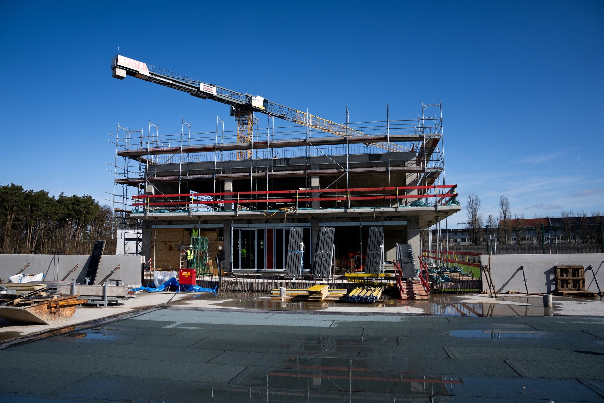 Ein im Bau befindliches Gebäude mit Gerüst und Kran, umgeben von Baumaterialien und einem klaren blauen Himmel.