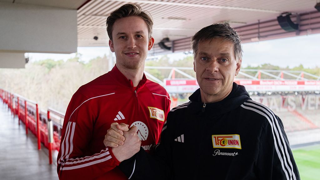 Zwei Männer in roten Fußballjacken stehen im Stadion, schütteln sich die Hände und lächeln in die Kamera.