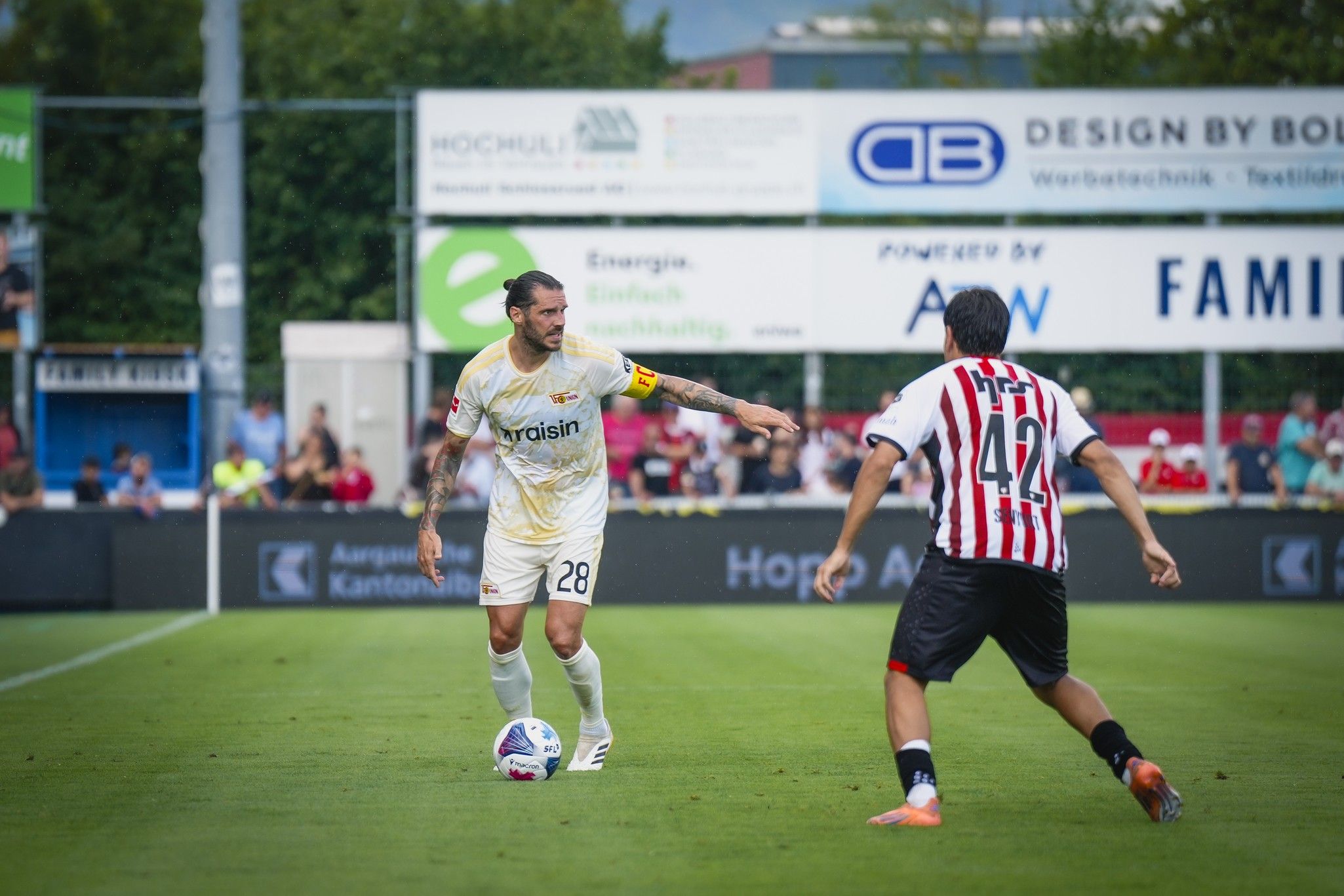 Ein Fußballspieler mit der Nummer 28 im gelben Trikot dribbelt den Ball, während ein Gegenspieler mit der Nummer 42 ihn verteidigt.