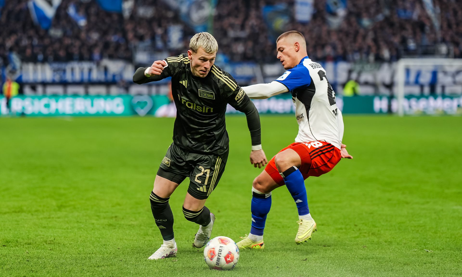 Zwei Fußballspieler im Wettkampf um den Ball auf einem Rasenfeld, Zuschauer im Hintergrund.