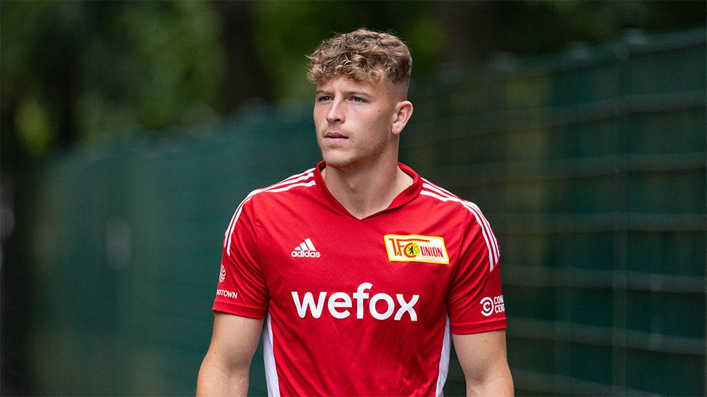 Ein junger Mann in einem roten Fußballtrikot mit dem Logo "wefox" läuft an einem grünen Zaun entlang.