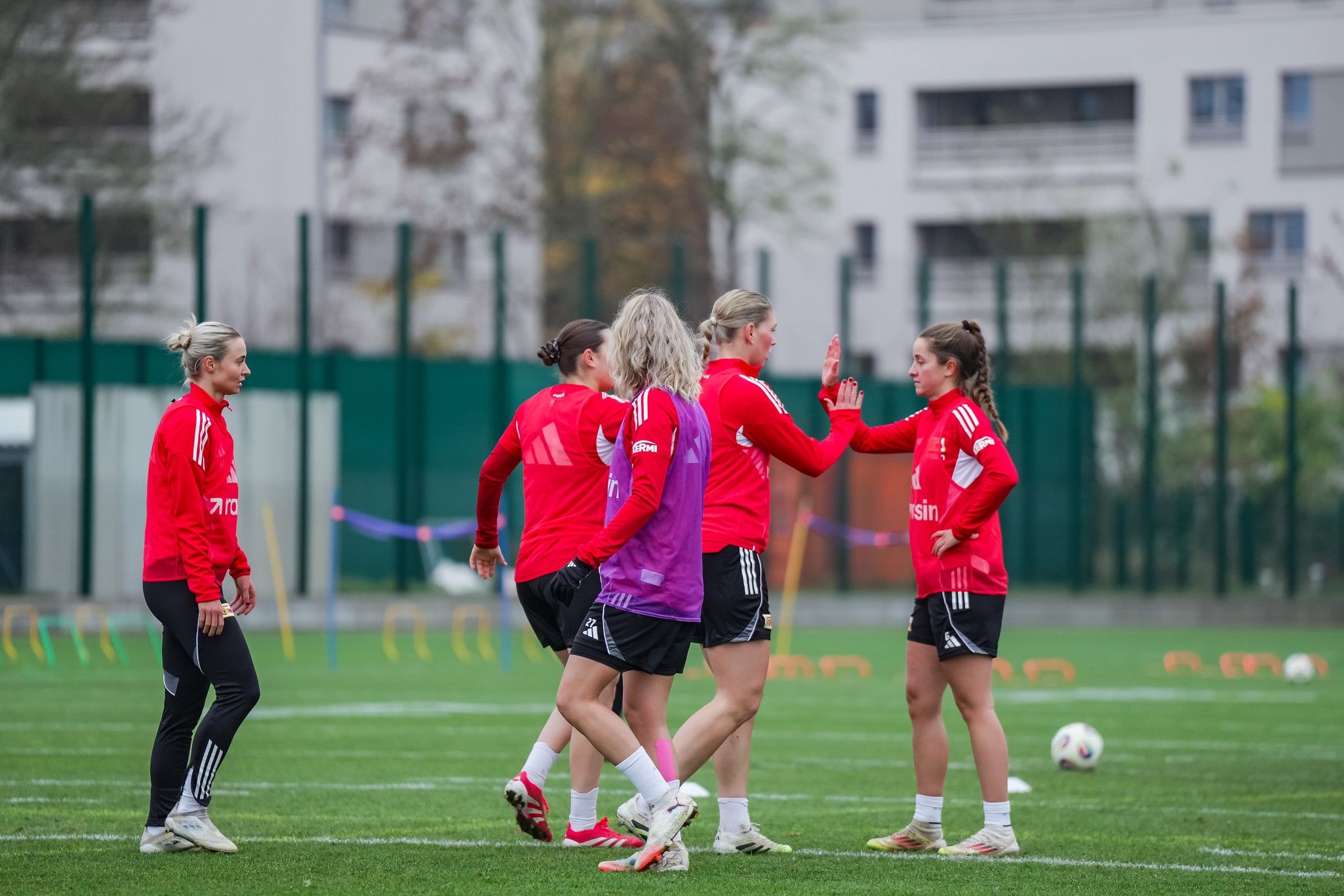 Frauenmannschaft beim Training auf einem Fußballplatz, in roten Trikots, während sie sich gegenseitig begrüßen.
