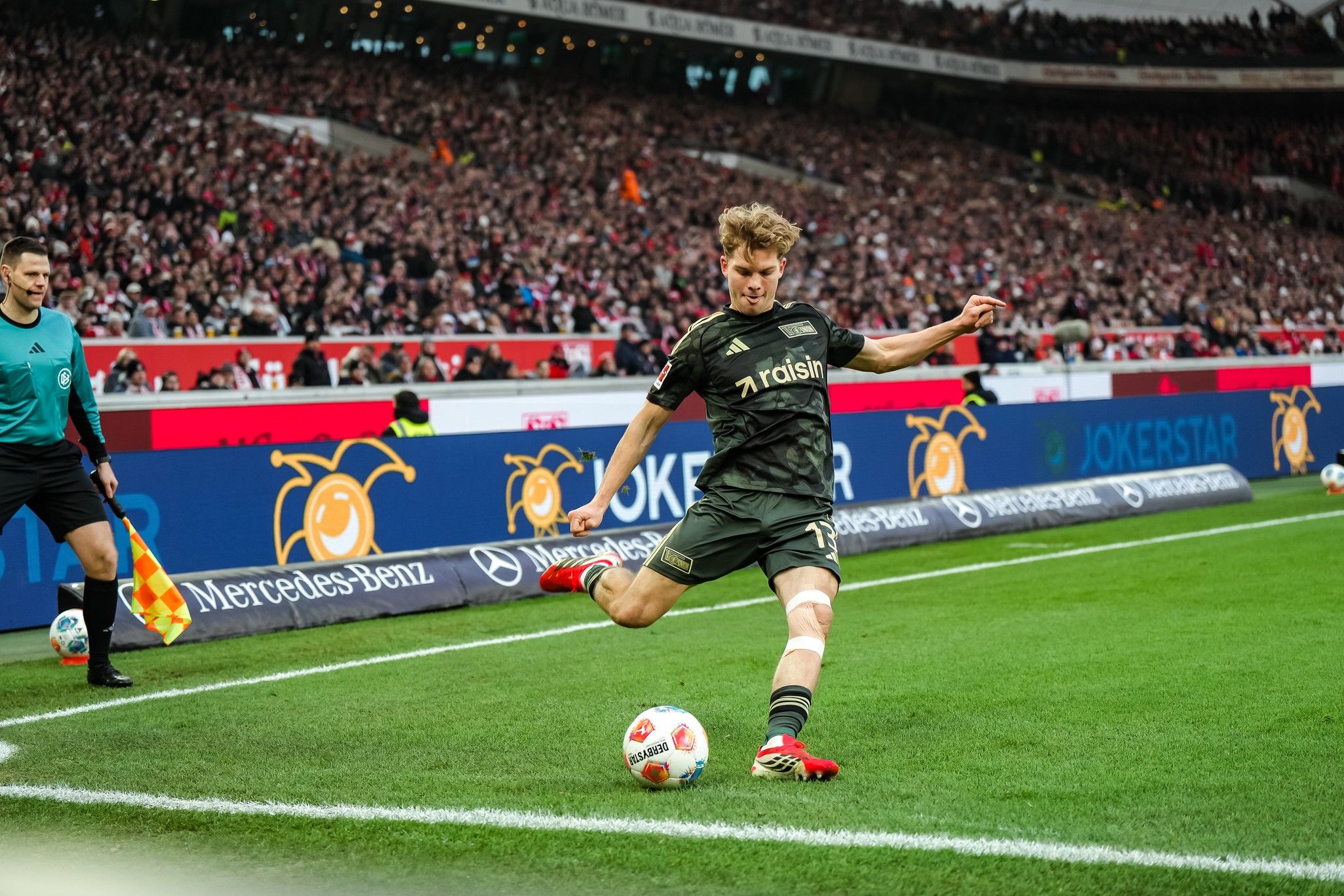 Fußballspieler in schwarzer Kleidung tritt den Ball an der Seitenlinie, Zuschauer im Hintergrund.