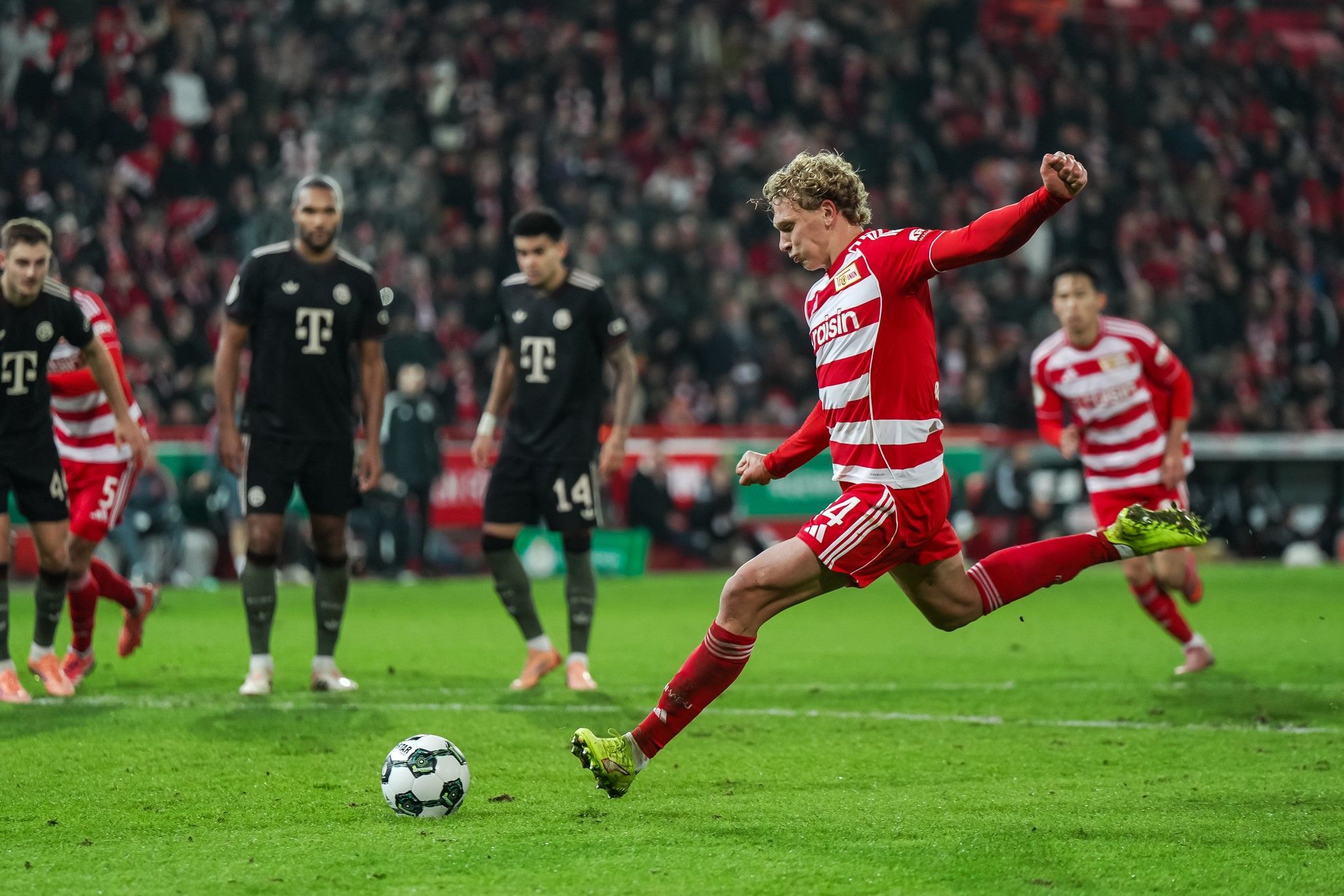 Ein Spieler in rot-gestreiftem Trikot führt einen Elfmeter aus, während Gegner in schwarzen Trikots zuschauen.