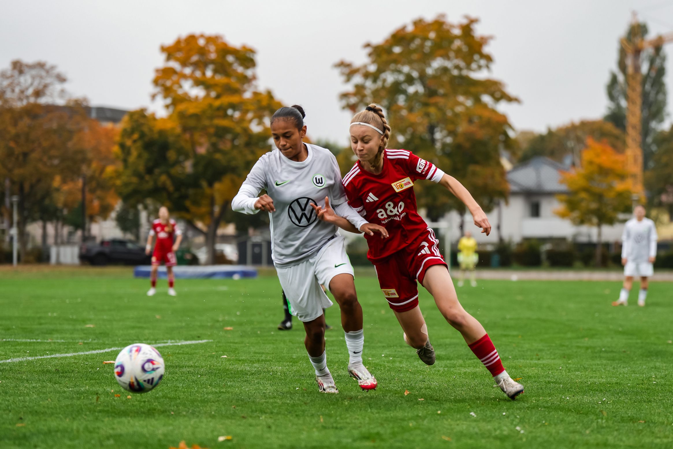 Zwei Fußballspielerinnen in Aktion auf einem Rasenplatz, umgeben von herbstlichen Bäumen.