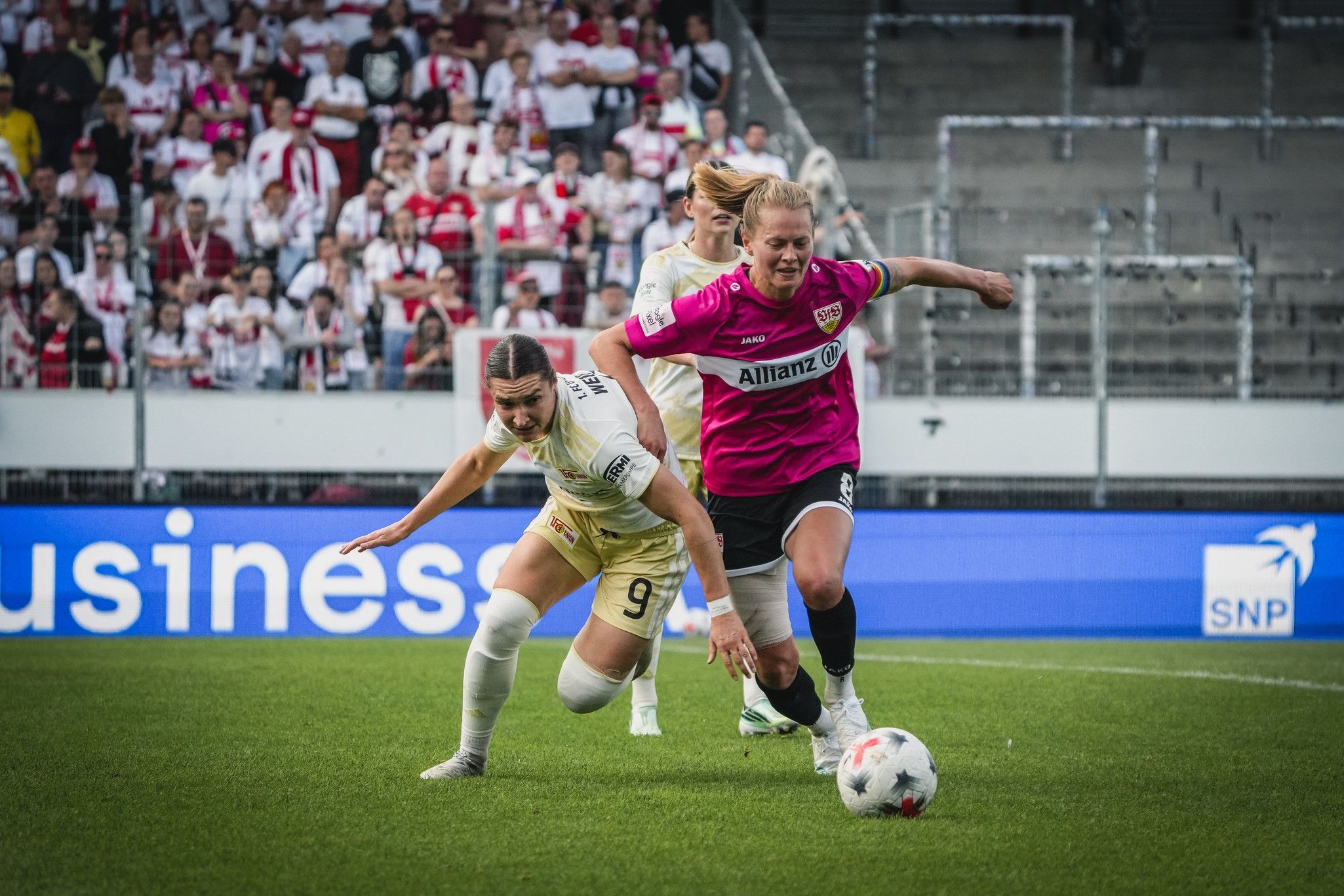 Zwei Fußballspielerinnen kämpfen um den Ball auf einem Spielfeld, während Zuschauer im Hintergrund jubeln.