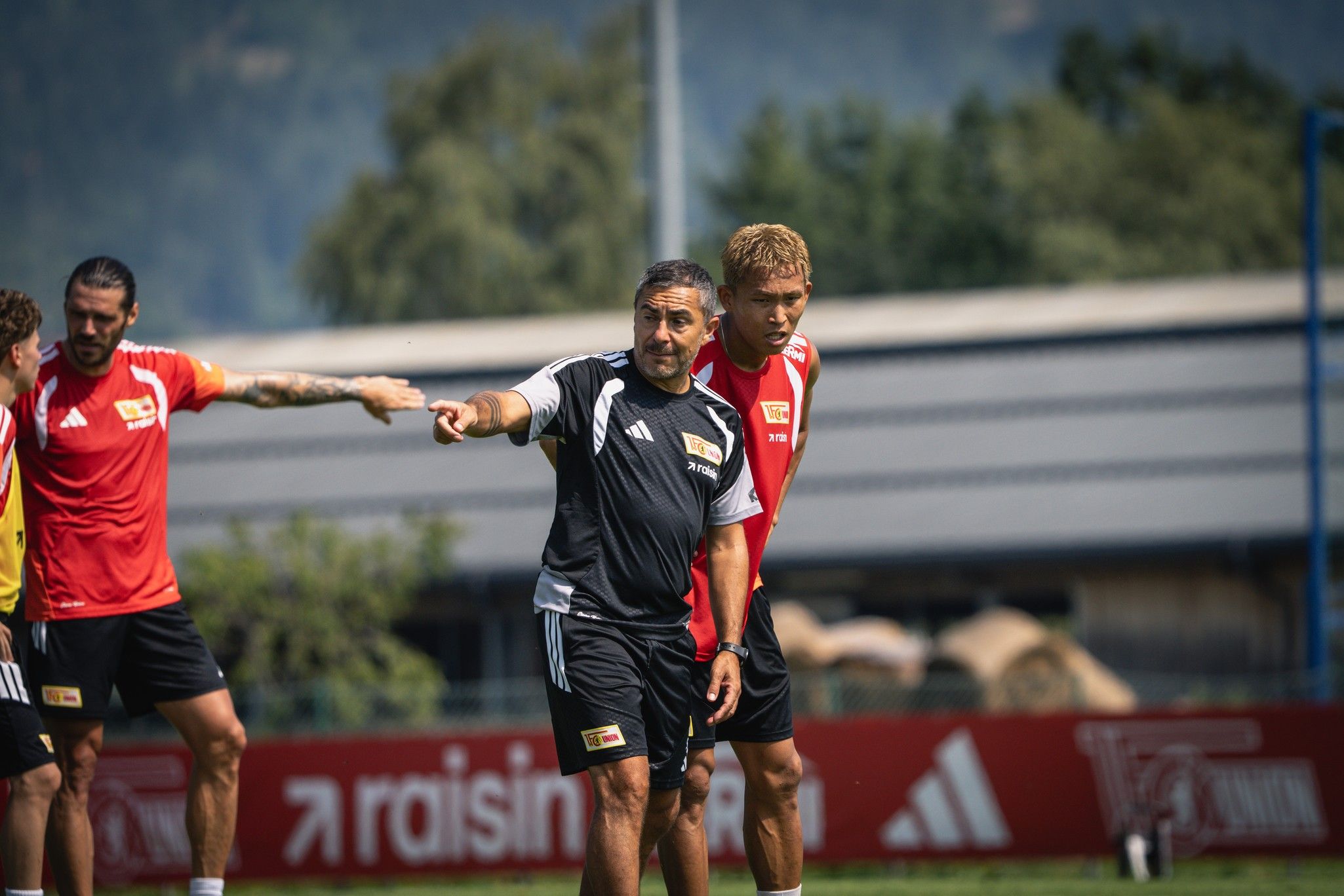 Trainer gibt Anweisungen während eines Fußballtrainings, Spieler hören aufmerksam zu, im Hintergrund Bäume und eine Sporteinrichtung.