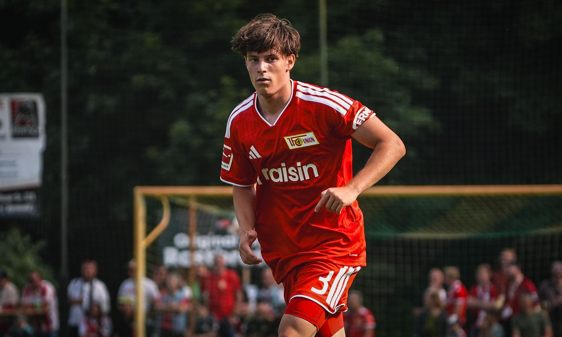 Ein junger Fußballspieler in rotem Trikot sprintet während eines Spiels auf dem Platz. Umgeben von Zuschauern im Hintergrund.