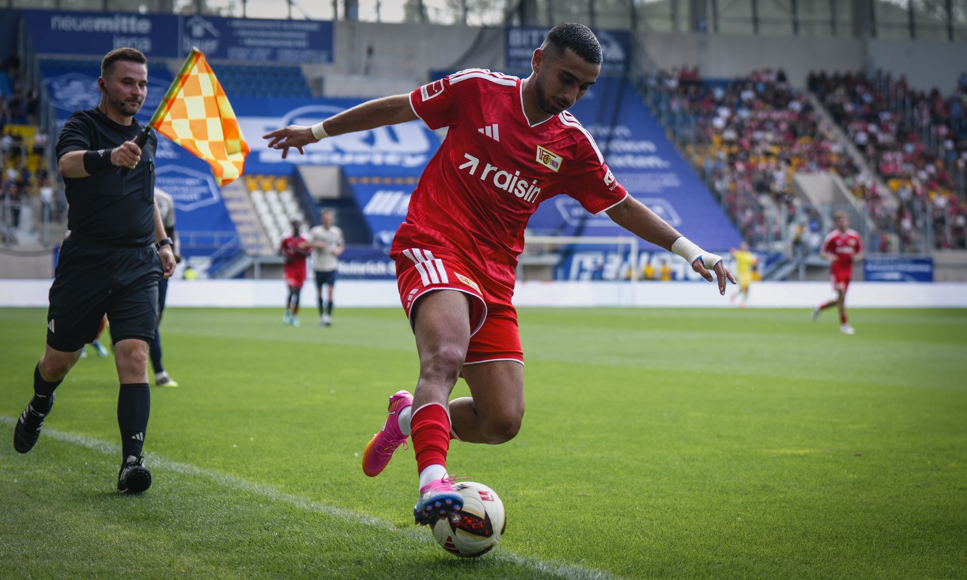 Spieler in rotem Trikot führt einen Einwurf an, während der Schiedsrichter im Hintergrund steht. Zuschauer im Stadion sind sichtbar.