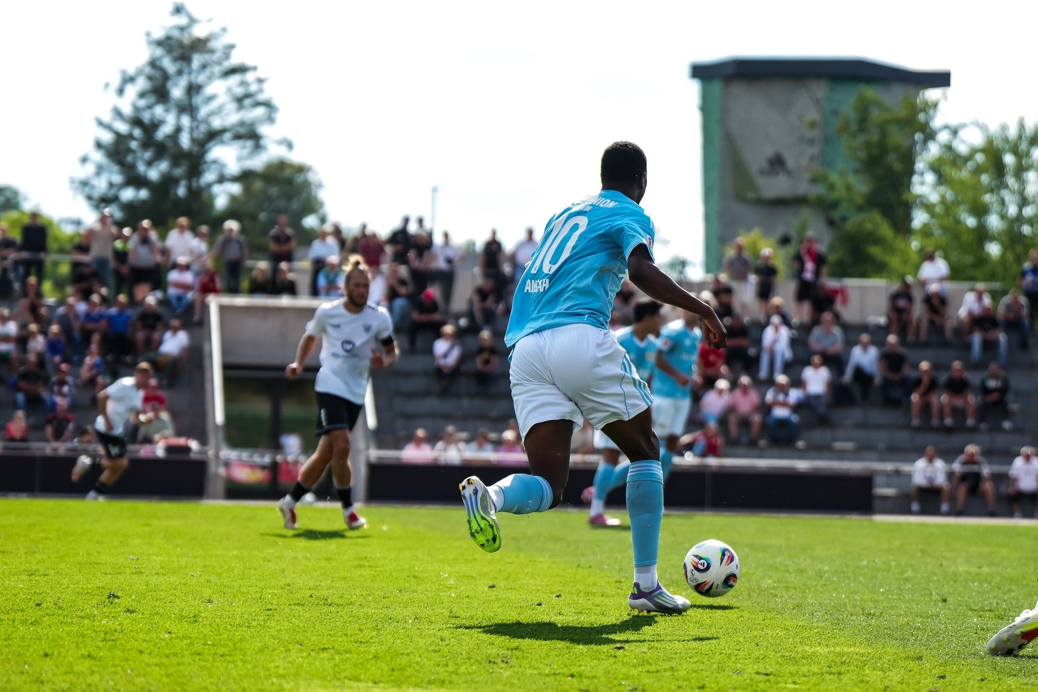 Ein Fußballspieler mit der Nummer 10 dribbelt den Ball auf einem grünen Feld, während Zuschauer im Hintergrund zuschauen.