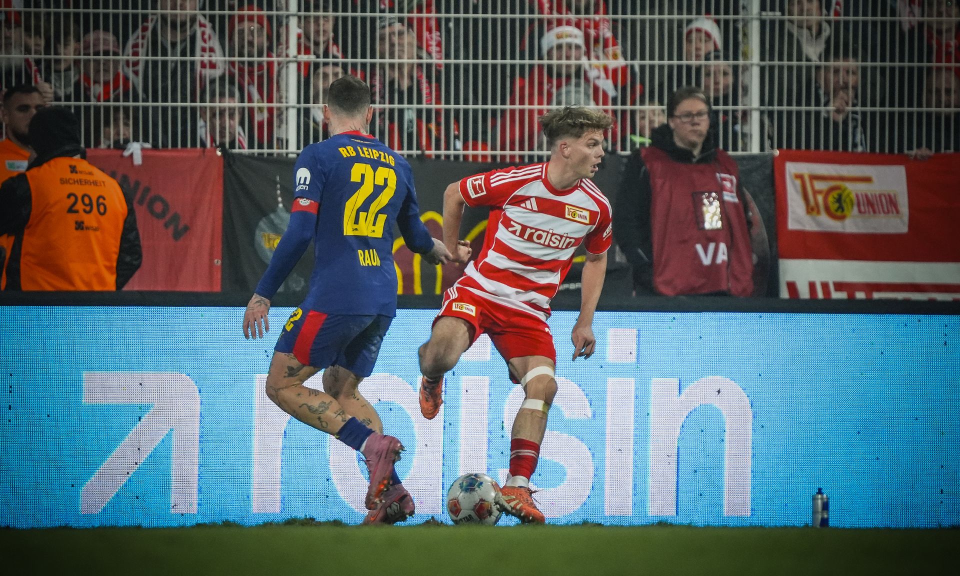 Ein Spieler in rot-weißer Uniform dribbelt den Ball, während ein Gegner in blau-gelber Uniform ihn verfolgt. Stadionhintergrund mit Fans.