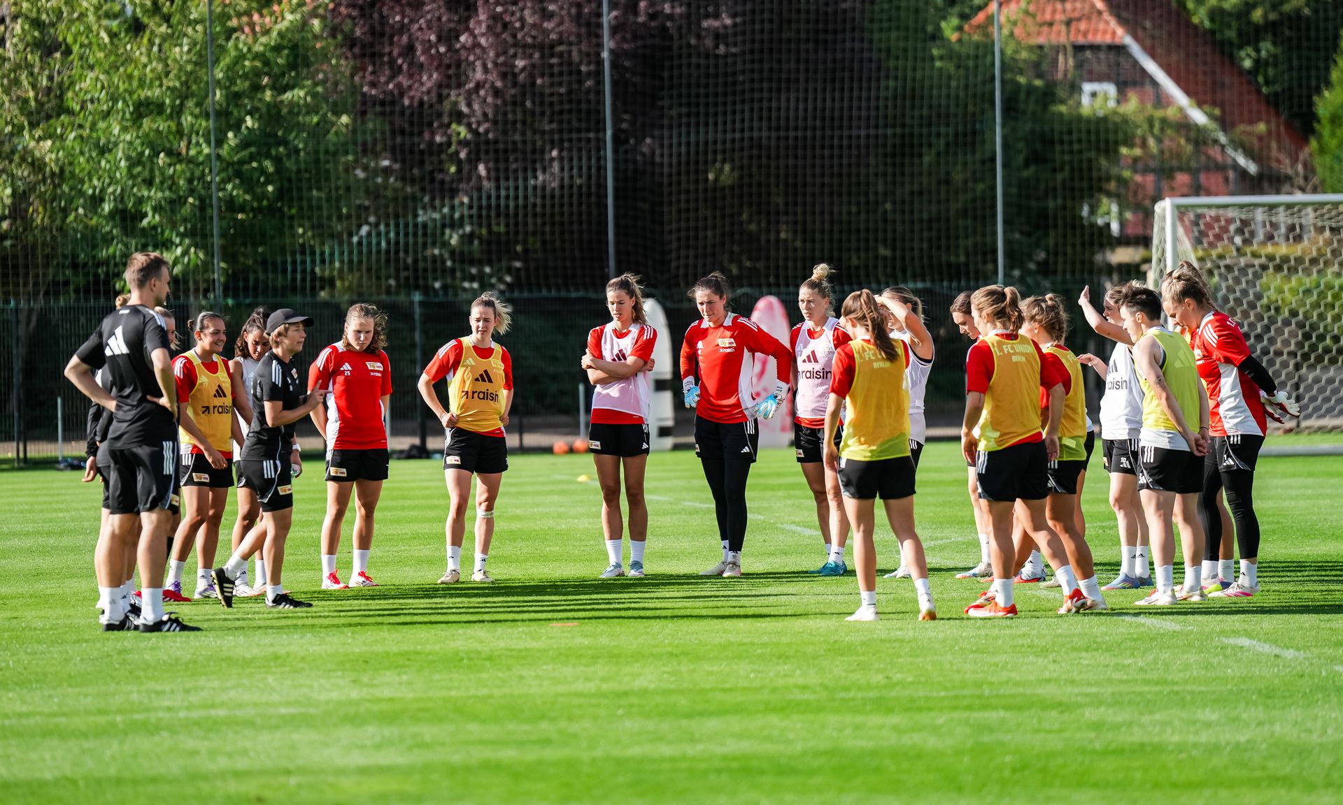Frauenfußballmannschaft versammelt sich um einen Trainer auf einem Trainingsplatz, um Anweisungen zu erhalten.