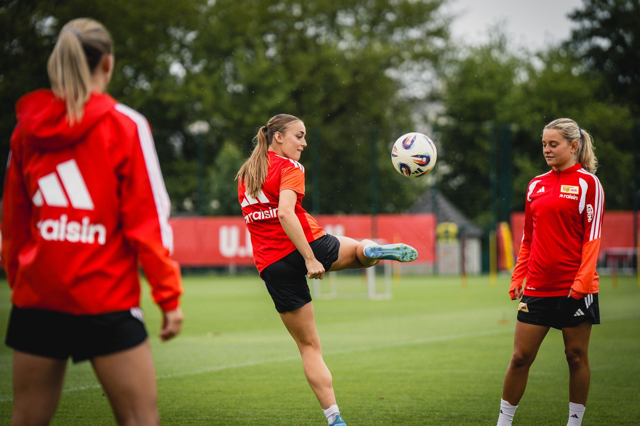 Drei Frauen in sportlicher Kleidung trainieren auf einem grünen Fußballplatz, während eine Spielerin den Ball hochhebt.
