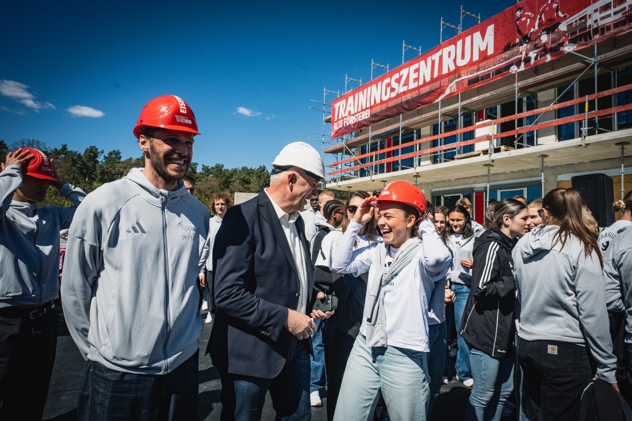 Personen in Arbeitskleidung und Helmen stehen vor einem Bauprojekt mit dem Schriftzug "TRAININGSSZENTRUM". Sonniger Himmel im Hintergrund.