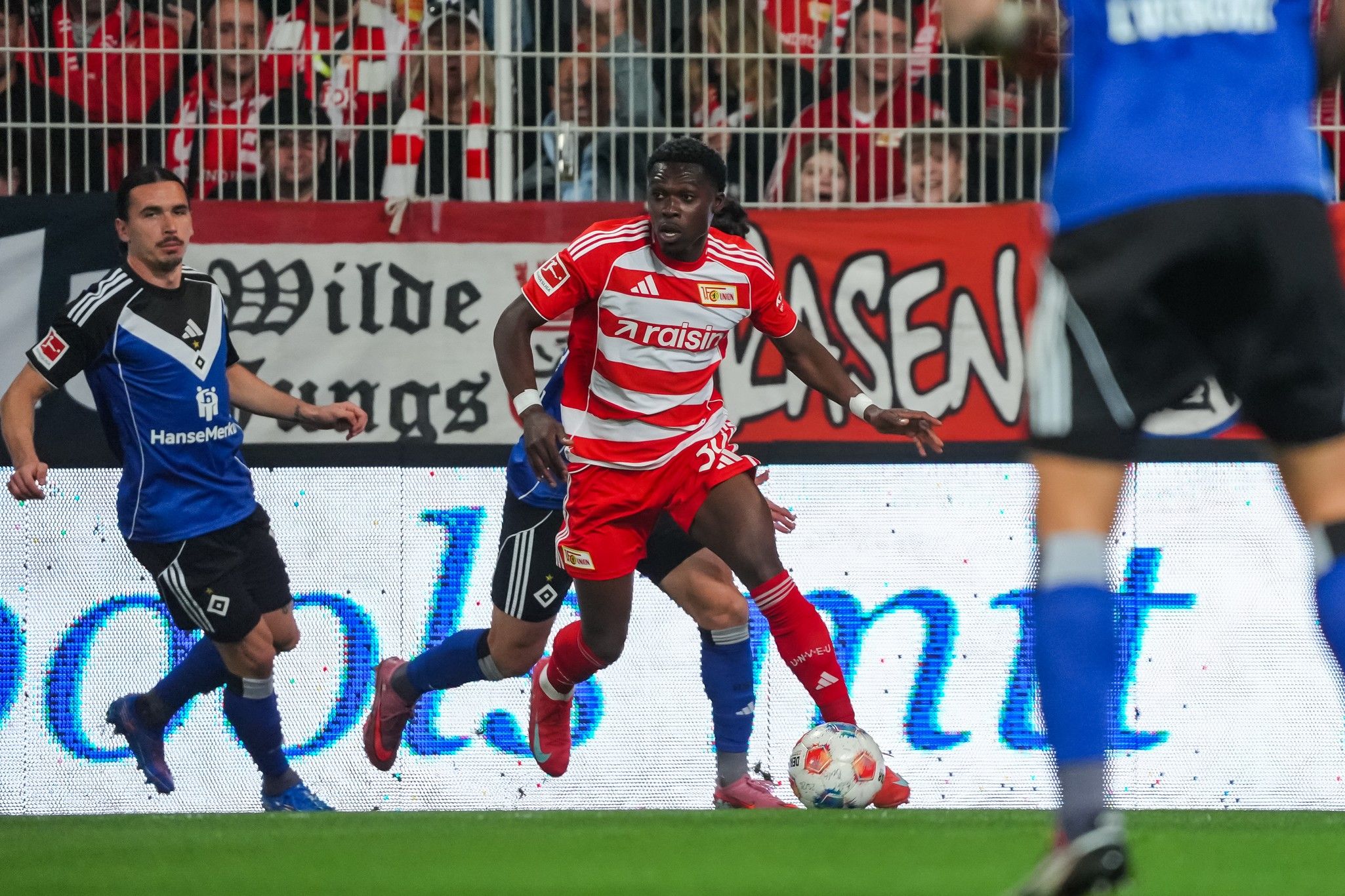 Ein Fußballspieler in rot-gestreifter Kleidung dribbelt den Ball, während zwei Gegenspieler in blauen Trikots versuchen, ihn zu stoppen.