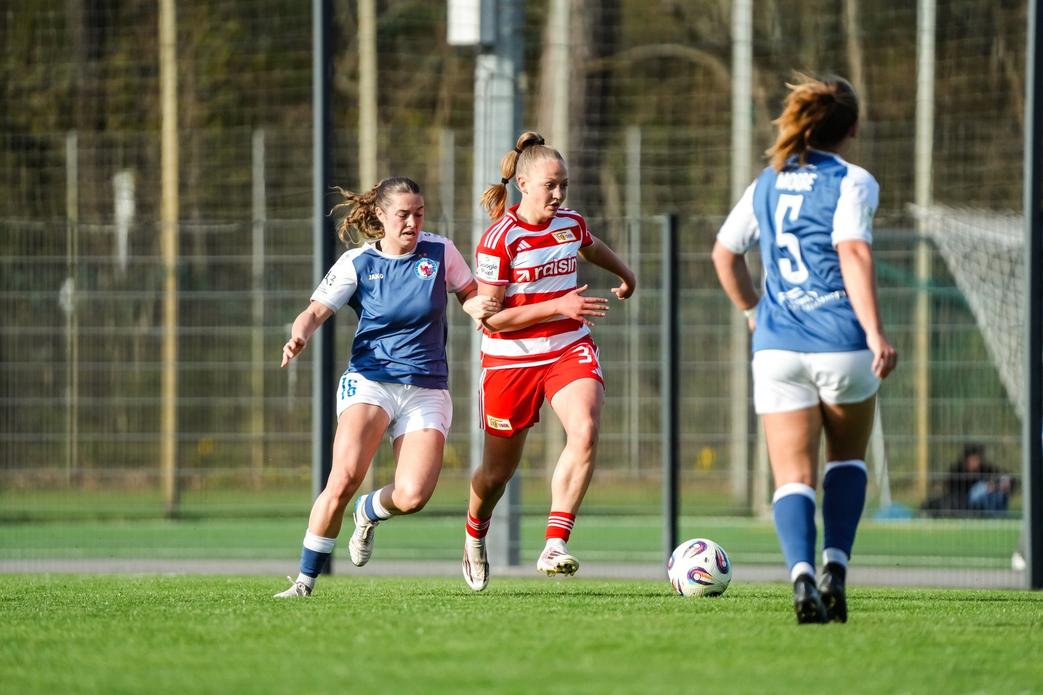 Zwei Fußballspielerinnen sprinten auf dem Spielfeld, während eine dritte Spielerin im Hintergrund steht.