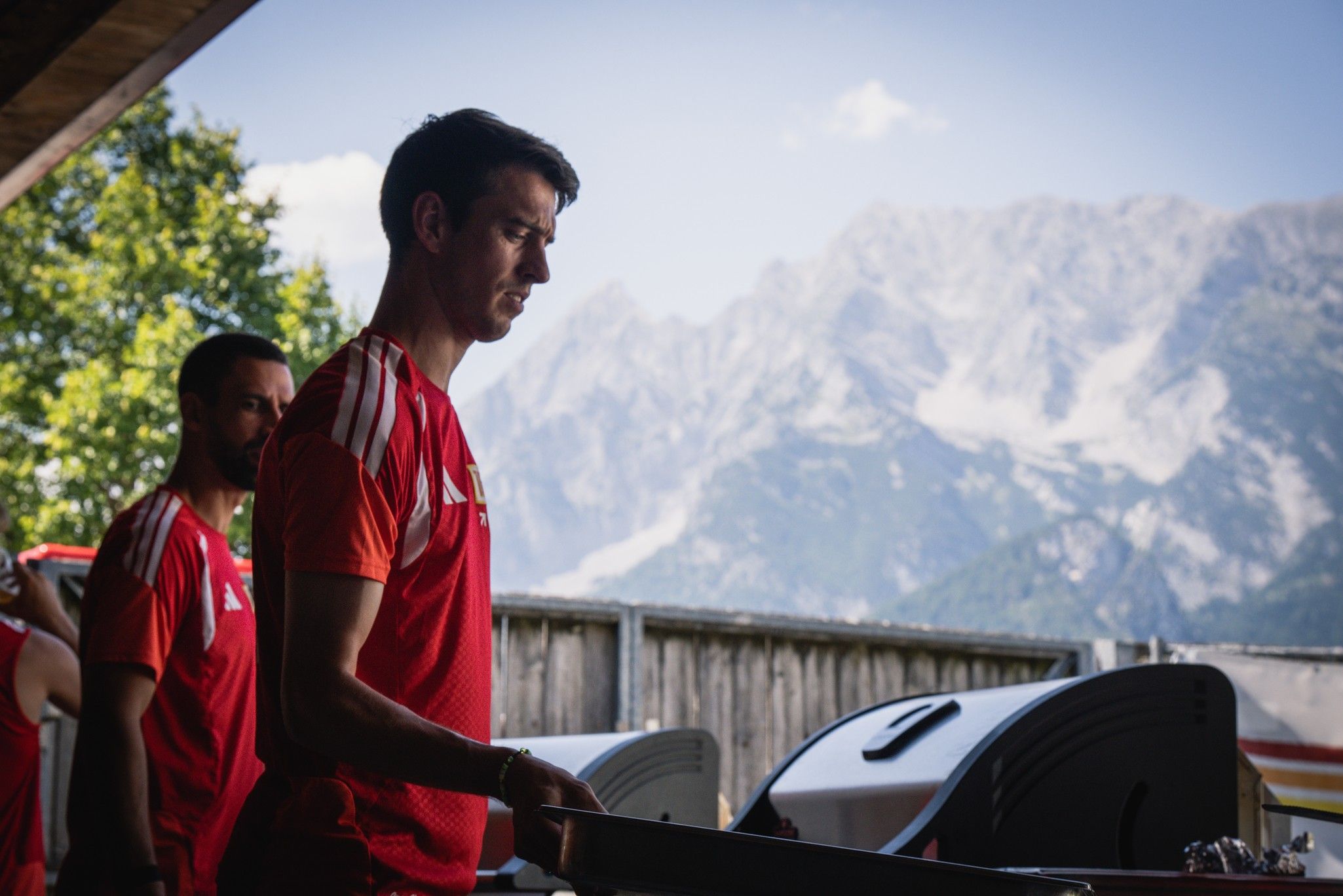 Ein Mann in rotem Trikot steht an einem Grill, im Hintergrund sind Berge und ein klarer Himmel zu sehen.