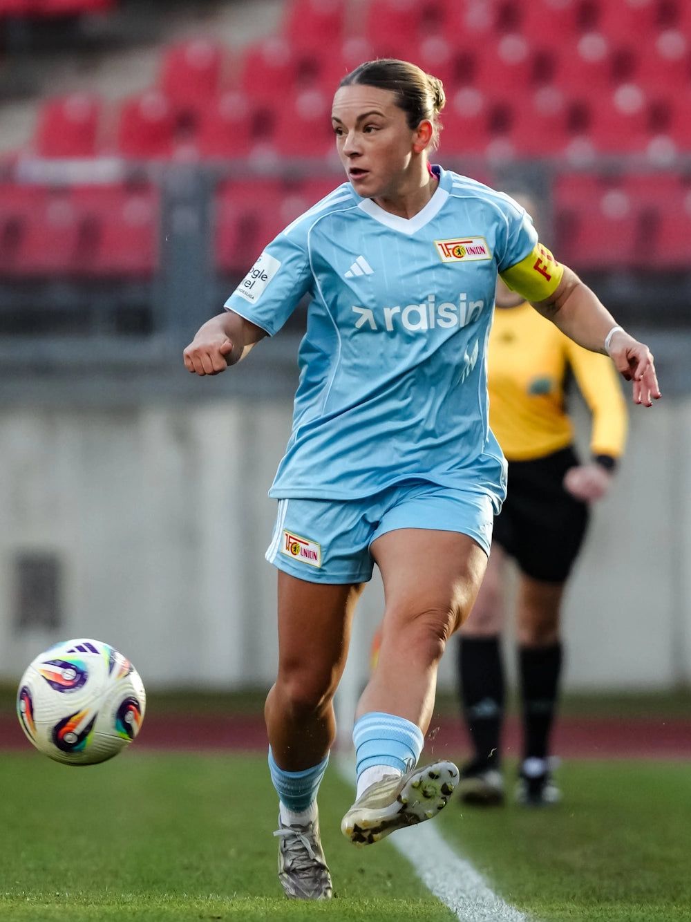 Frauenfußballspielerin in himmelblauer Uniform, dribbelt den Ball auf dem Platz. Im Hintergrund ein Schiedsrichter.