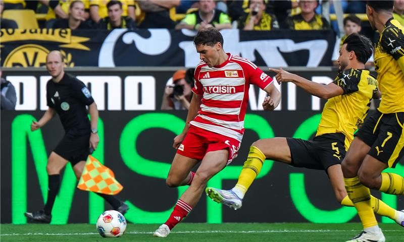 Ein Fußballspieler in rot-weißer Uniform schießt den Ball, während zwei Spieler in gelb-schwarzen Trikots ihn verteidigen.